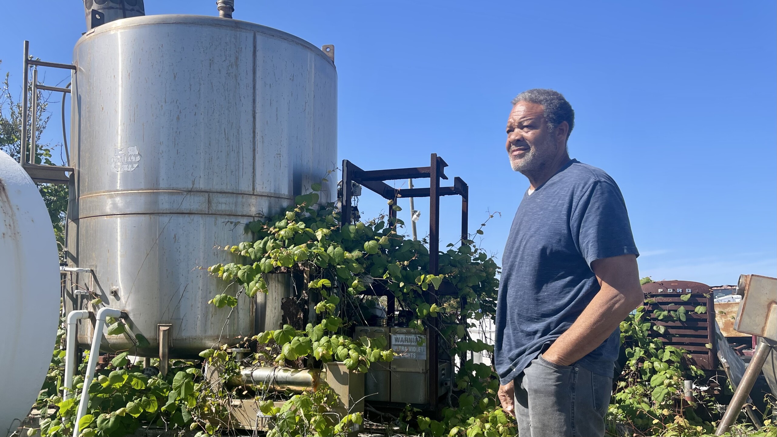 An older African-American man, Bobby Dollison, stands in front of two large steel barrels with green shrubs growing around them on American Beach.