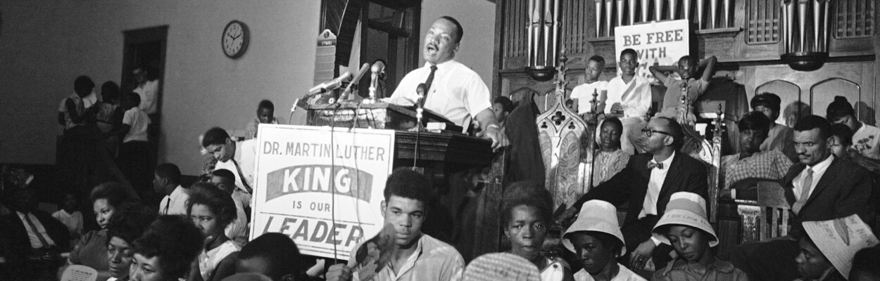 The Rev. Martin Luther King Jr. speaks at a lectern surrounded bya crowd.