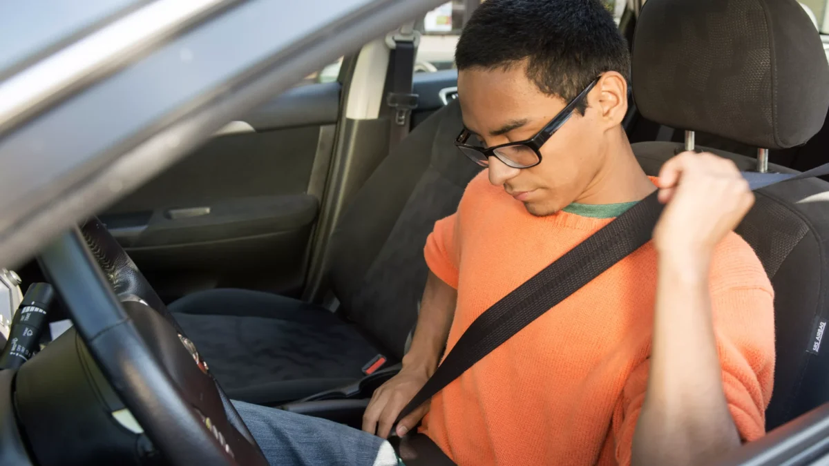 A young man in an orange shirt sits in the drivers side of a vehicle and buckles his seatbelt.