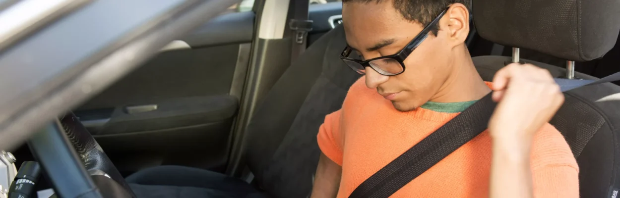 A young man in an orange shirt sits in the drivers side of a vehicle and buckles his seatbelt.