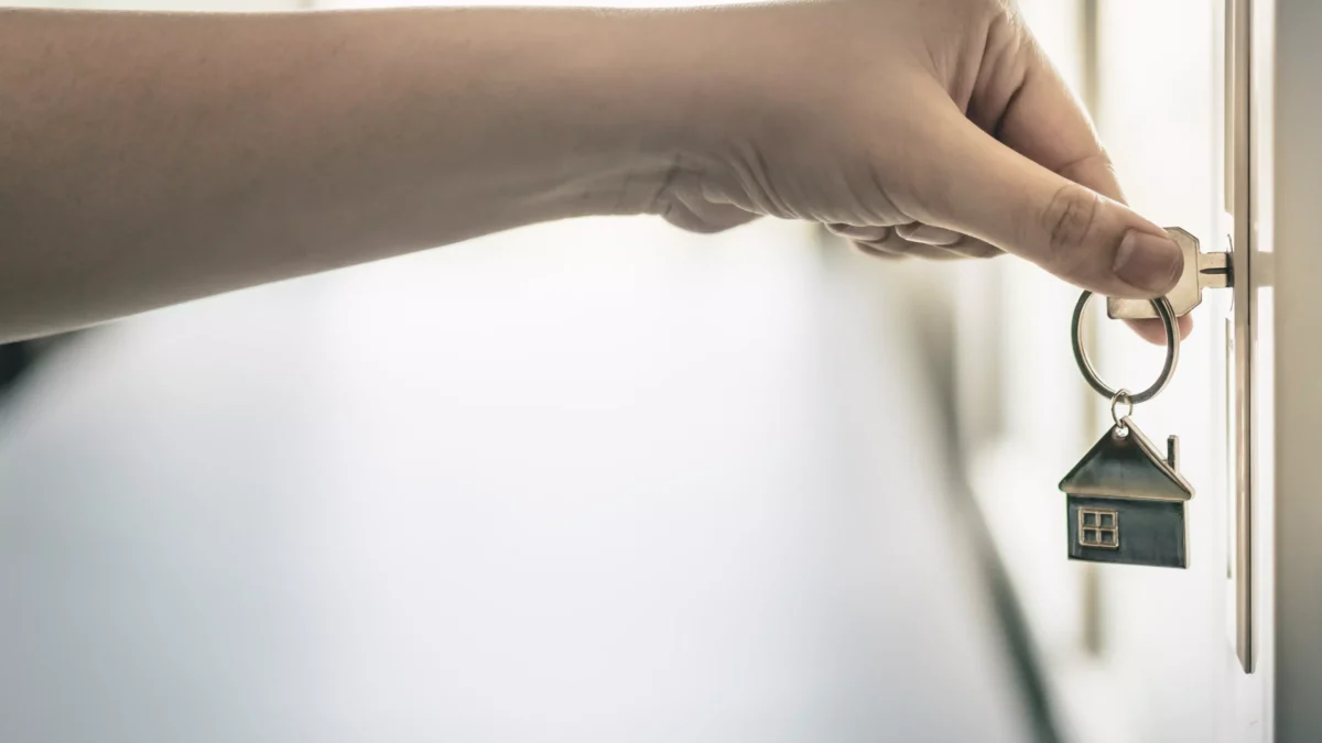 An arm holding a key with a house-shaped charm turns a key in the door of a house.