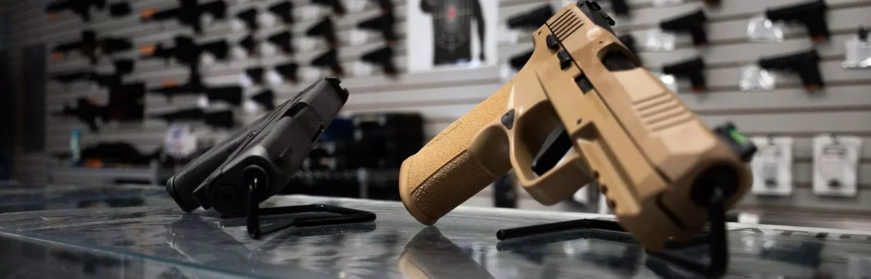 Two hand guns on top of a glass display. In the background are guns mounted on a wall.