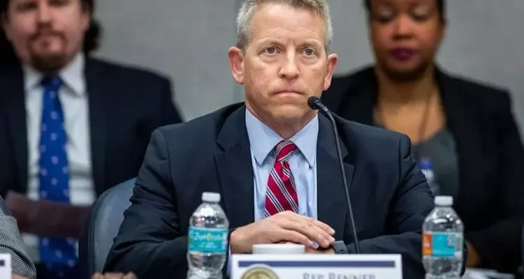 A stern-faced man wearing a dark blue suit with a red striped tie sits with his arms folded across a table. He's facing a microphone.