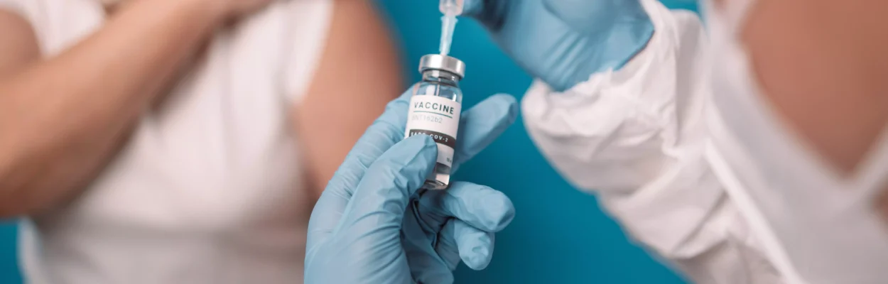 a health provider prepares a covid shot while in the blurred background a patient rolls up sleeve awaiting to receive the vaccine