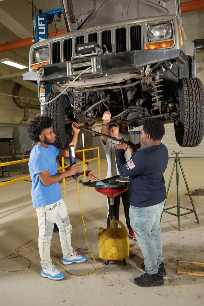 Three CTE students in an automotive shop stand below a Jeep on a lift