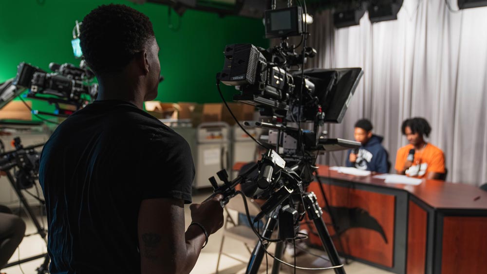 Three CTE students in a TV studio - one behind a camera and two at a news desk