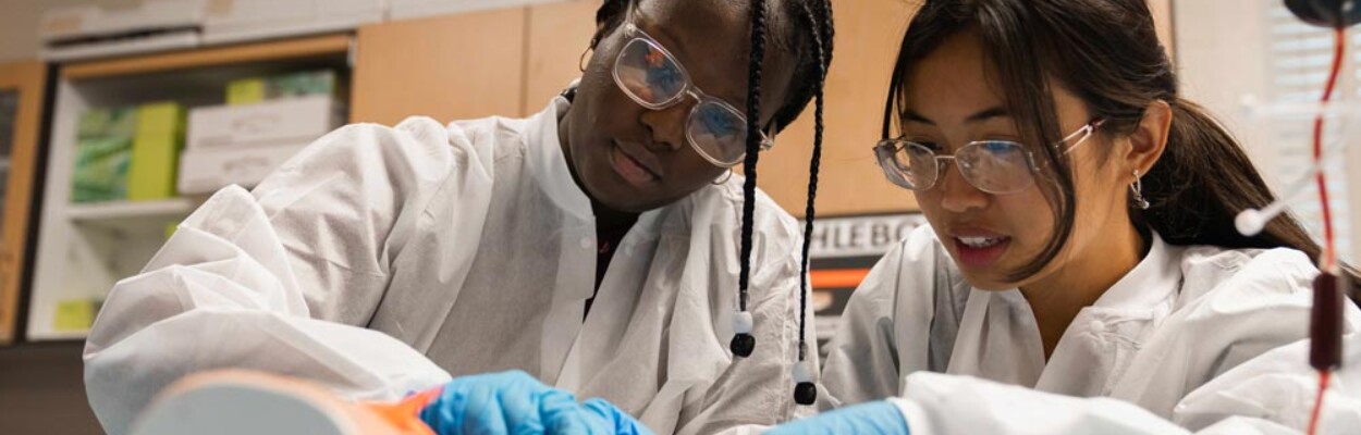 Two female career and technical education students wear lab coats and gloves while working on a simulated medical procedure.