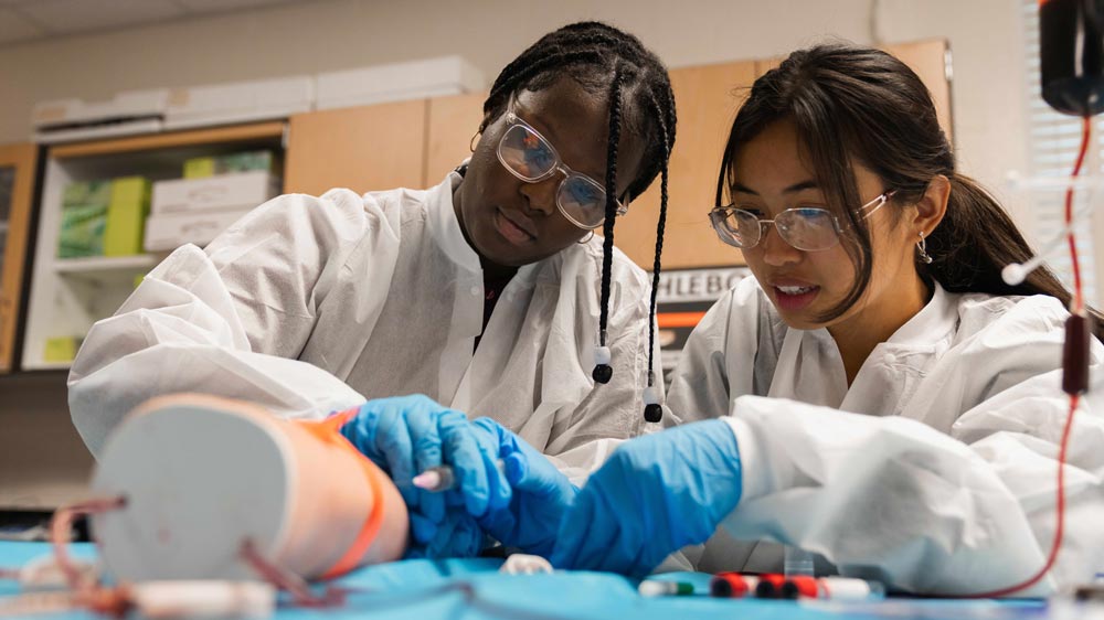 Two female career and technical education students wear lab coats and gloves while working on a simulated medical procedure.