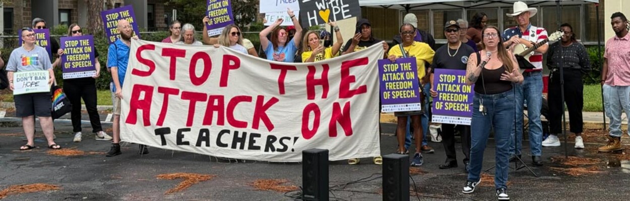 Protesters stand in front of DCPS headquarters holding a large banner that reads "STOP THE ATTACK ON TEACHERS" in bold, handwritten letters. The protest was in support of teachers' right to free speech.