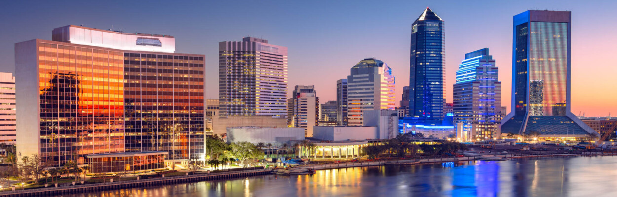 Skyline of Jacksonville, Florida with modern skyscrapers reflecting in the St. Johns River. Dawn light creates a gradient from deep blue to orange in the sky, casting reflections on the building facades and water. Prominent structures include mirrored glass and unique high-rise architectural designs. The water appears calm, enhancing the reflections and adding a serene ambiance to the bustling urban scene below.