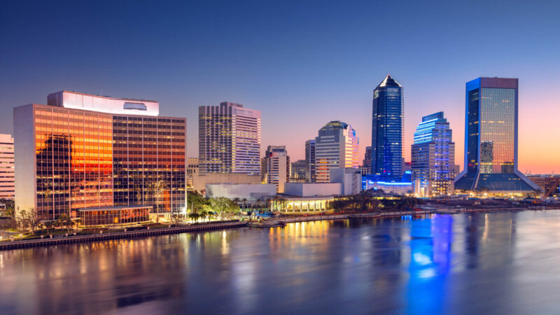 Skyline of Jacksonville, Florida with modern skyscrapers reflecting in the St. Johns River. Dawn light creates a gradient from deep blue to orange in the sky, casting reflections on the building facades and water. Prominent structures include mirrored glass and unique high-rise architectural designs. The water appears calm, enhancing the reflections and adding a serene ambiance to the bustling urban scene below.