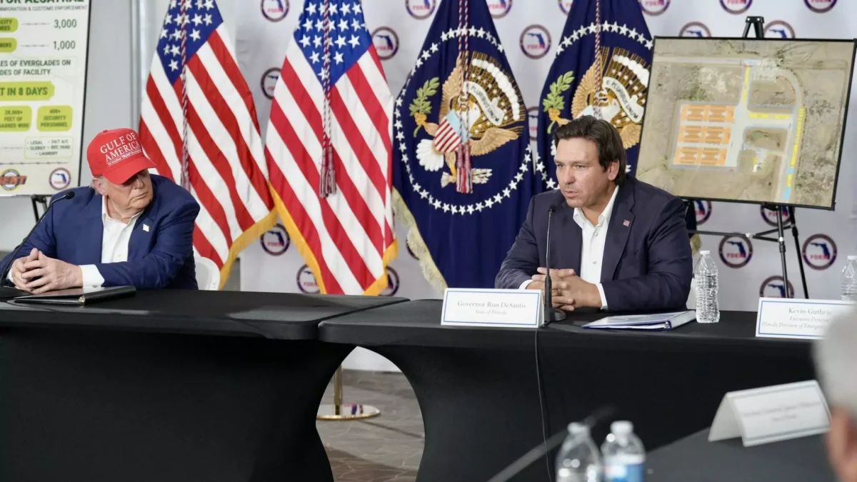 President Donald Trump and Florida Gov. Ron DeSantis sit at a table speaking to people. Flags are in the background.