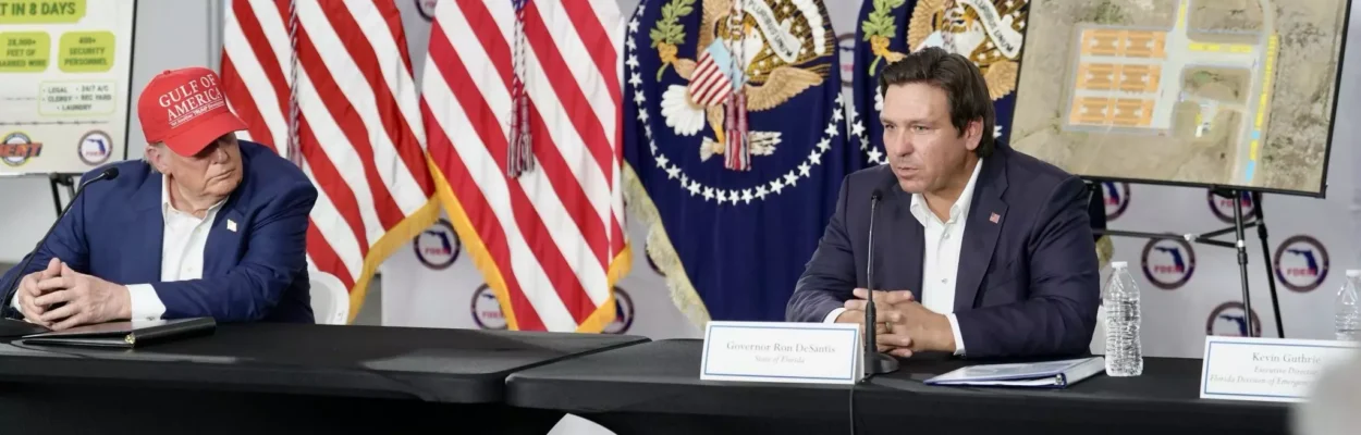 President Donald Trump and Florida Gov. Ron DeSantis sit at a table speaking to people. Flags are in the background.