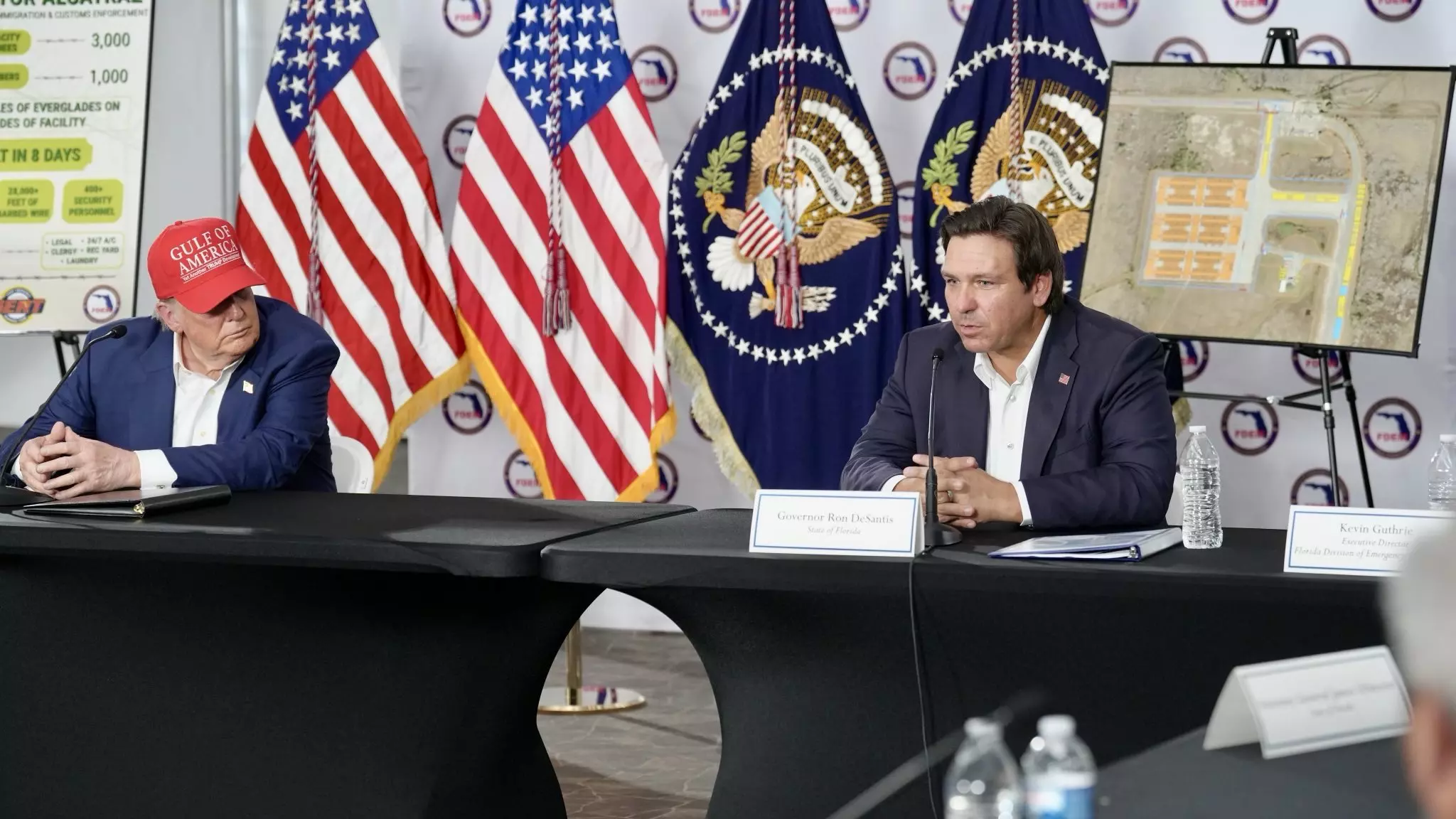 President Donald Trump and Florida Gov. Ron DeSantis sit at a table speaking to people. Flags are in the background.
