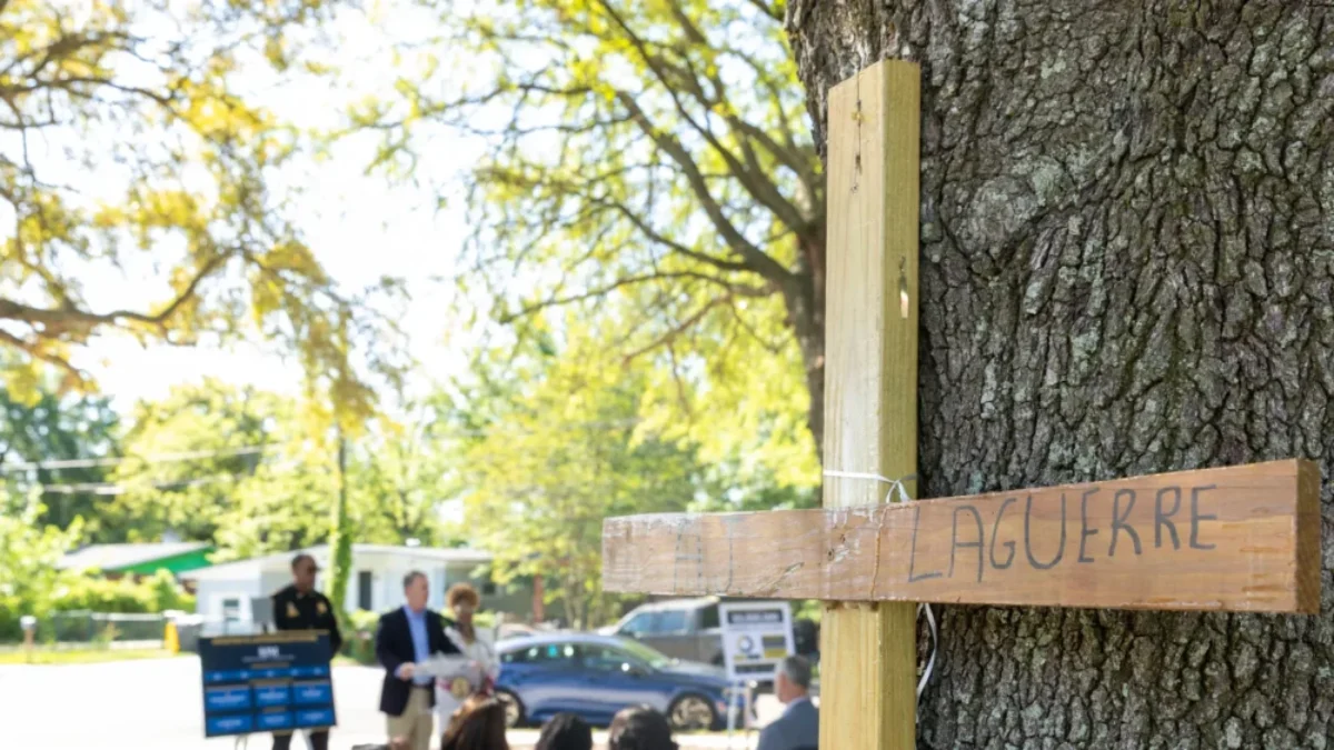 wooden cross memorial inscribed with a name, against a tree in the Northwest Jacksonville park where the press conference is seen behind.