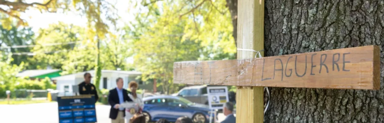 wooden cross memorial inscribed with a name, against a tree in the Northwest Jacksonville park where the press conference is seen behind.