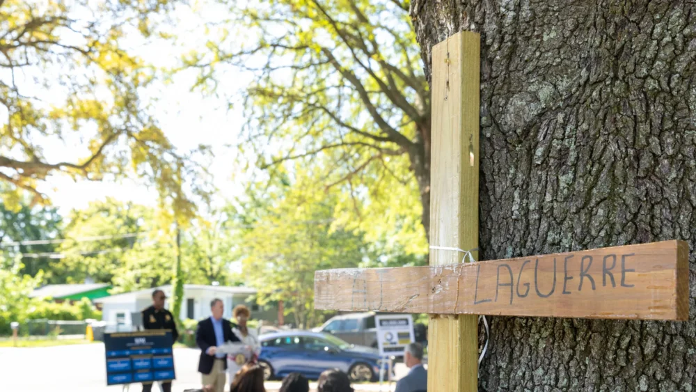 wooden cross memorial inscribed with a name, against a tree in the Northwest Jacksonville park where the press conference is seen behind.