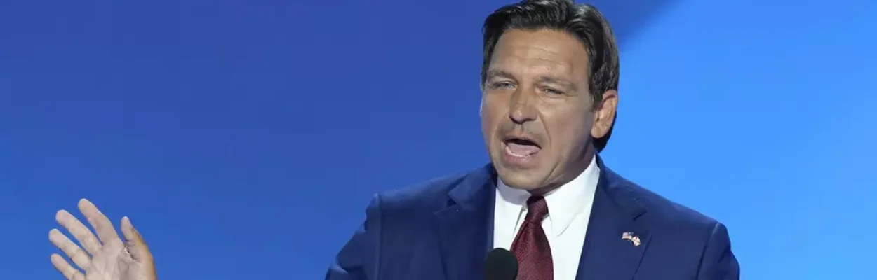 A man with brown hair wearing a blue suit and red tie speaking behind a microphone with his right hand raised in the air.