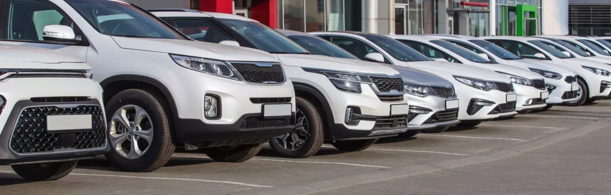 A row of new cars line the lot at a dealership, a row of white and silver vehicles in front of a building with many broad windows.