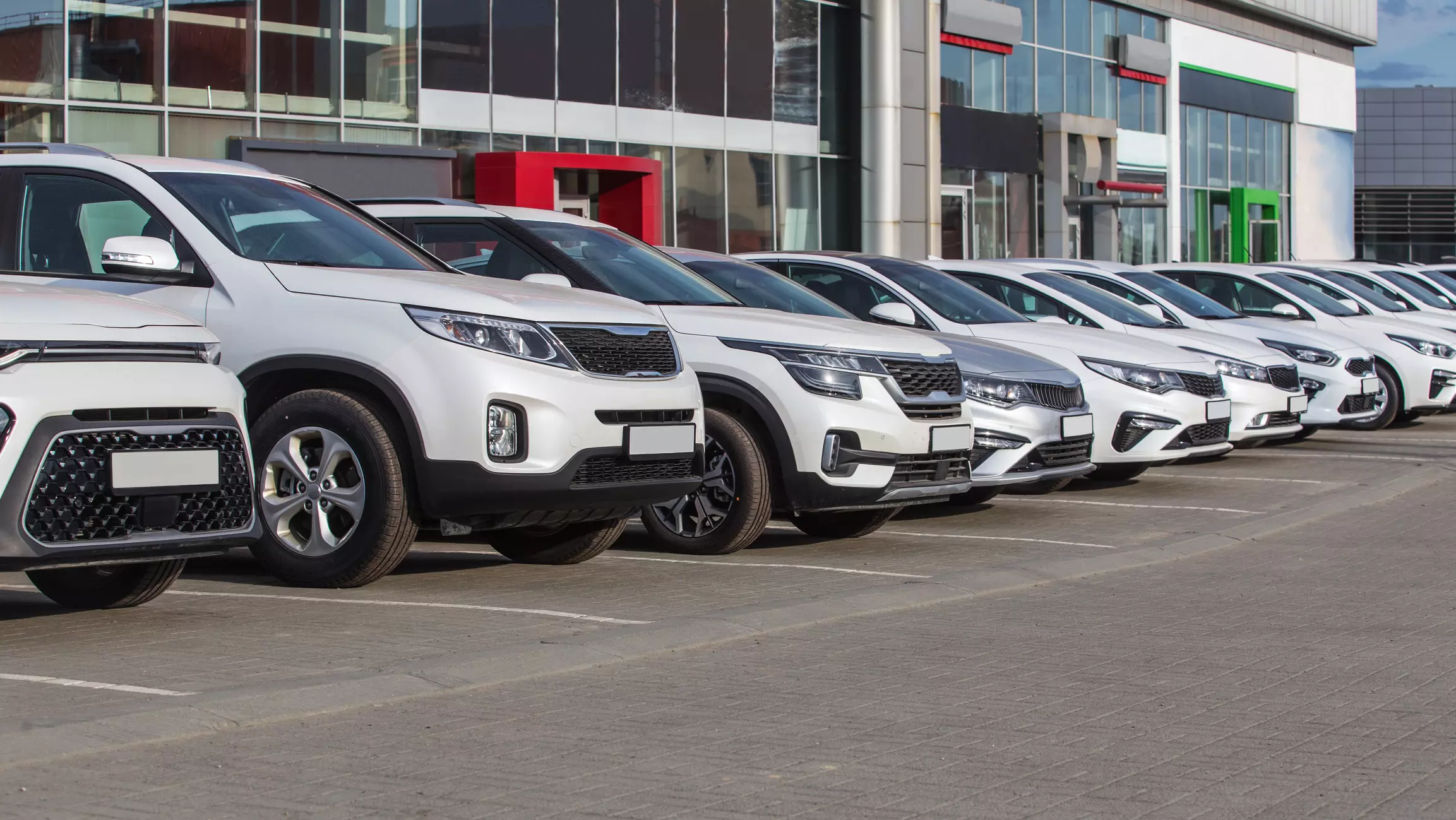 A row of new cars line the lot at a dealership, a row of white and silver vehicles in front of a building with many broad windows.