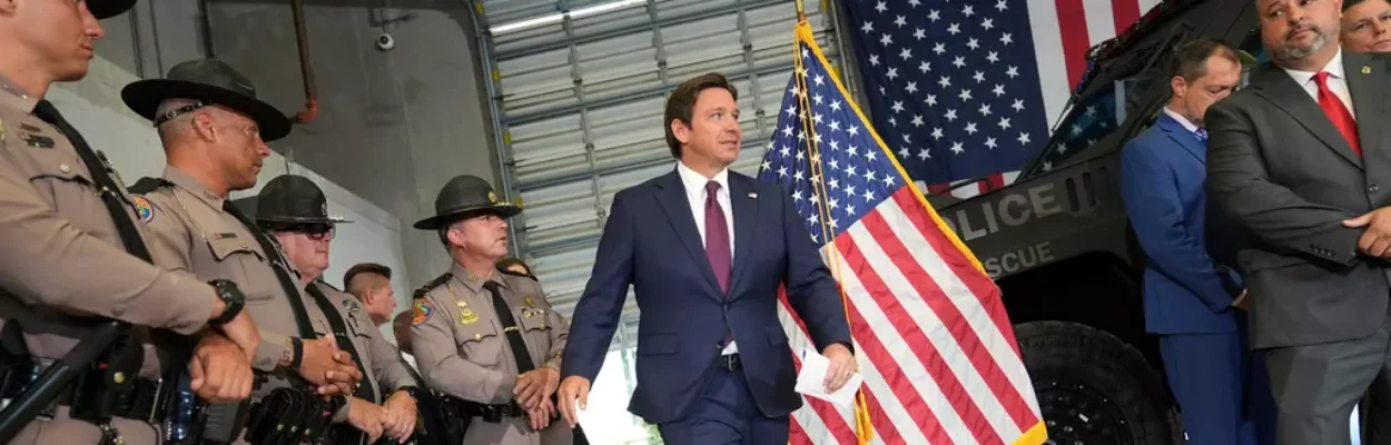 Man with brown hair wearing a blue suit is walking inside a building past a US flag during an immigration press conference. To the left are men in police uniforms, and a police vehicle in the background