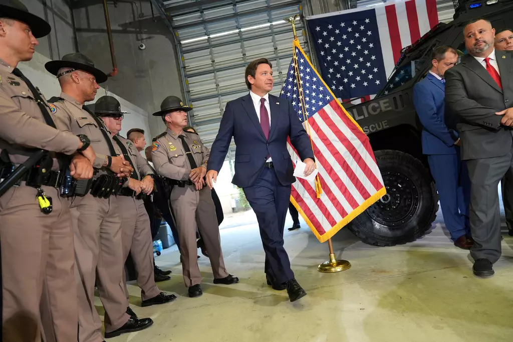 Man with brown hair wearing a blue suit is walking inside a building past a US flag during an immigration press conference. To the left are men in police uniforms, and a police vehicle in the background