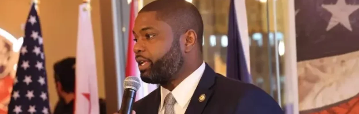 Black man with a beard and dark suit speaking into the microphone, looking to the left and an American flag to the left behind him