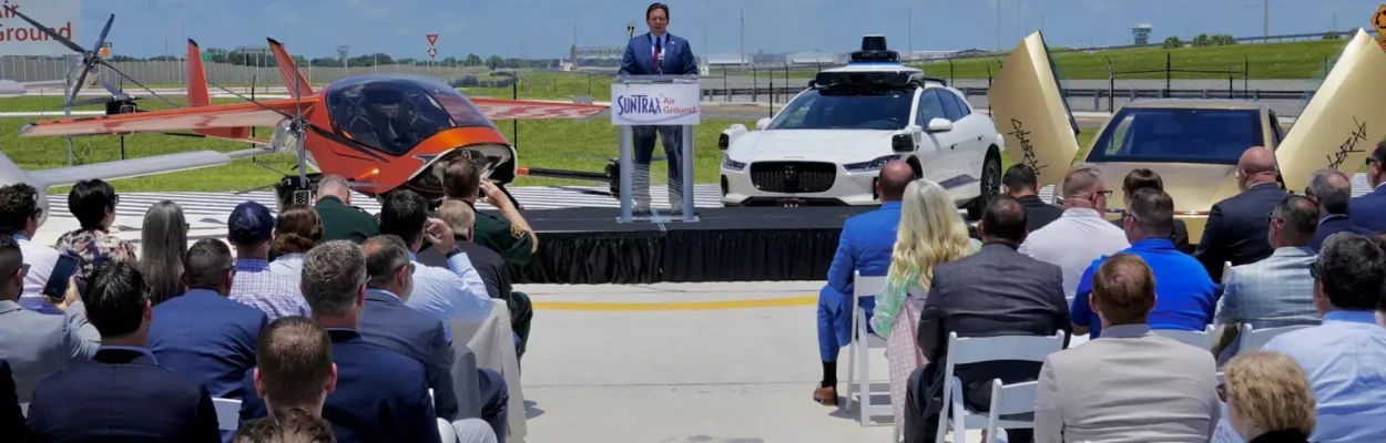 Man on podium with cars and an eVTOL behind him. Crowd of people sit in white chairs in front of him.