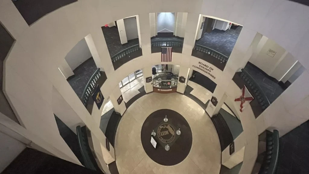 A view looking down through the open rotunda of the Leon County Courthouse in Tallahassee. On the ground floor, the county seal is embedded in the tile floor beneath an American flag hanging from the second level.