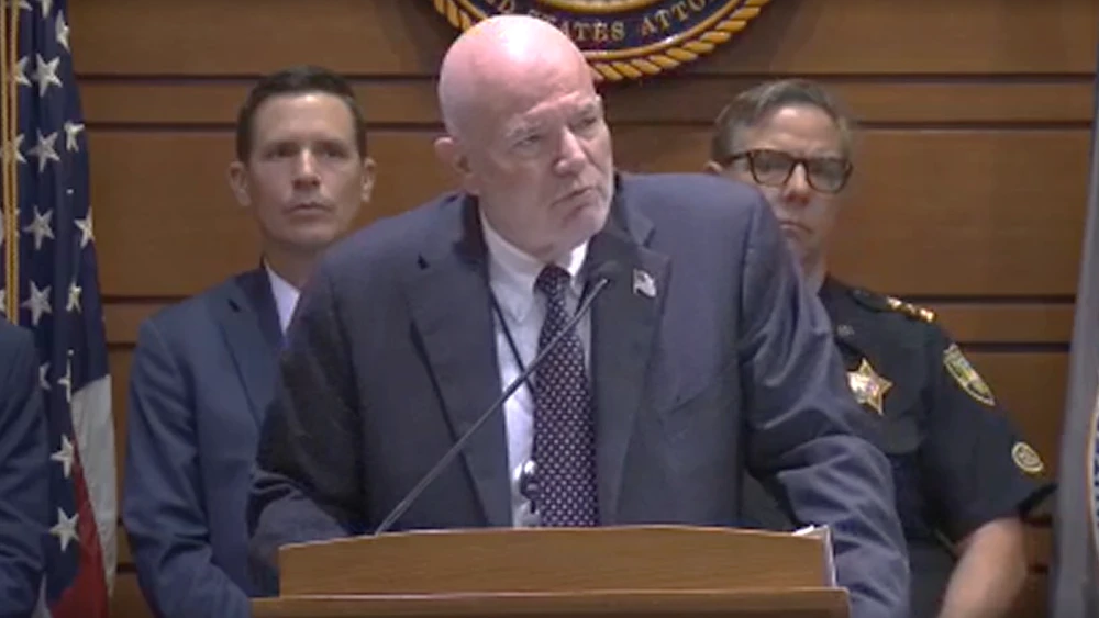 U.S. Attorney Gregory Kehoe, wearing a gray suit, stands in front of a microphone discussing gang arrests.