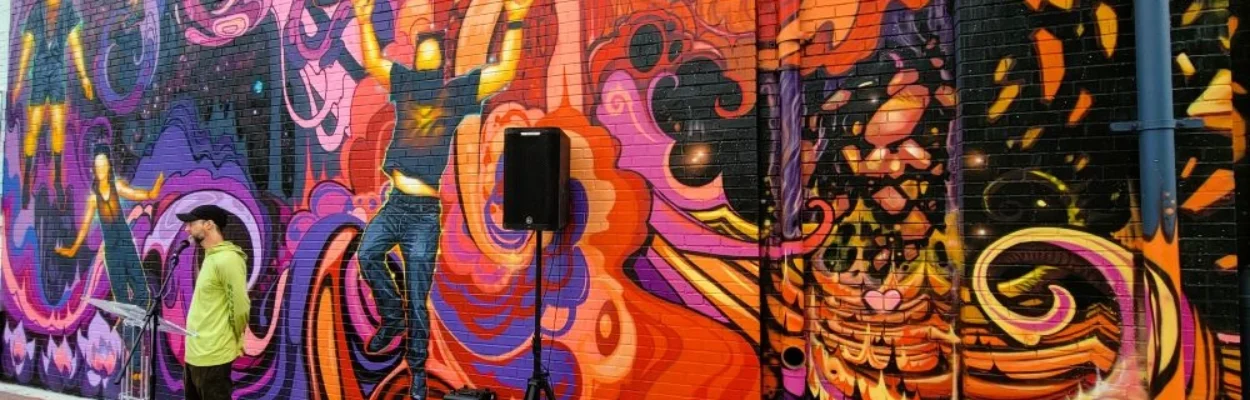 A man in a ballcap stands at a microphone in front the the vibrant colors of a new Five Points mural.