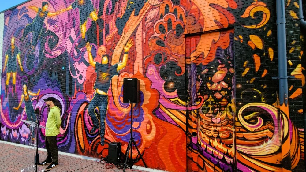 A man in a ballcap stands at a microphone in front the the vibrant colors of a new Five Points mural.