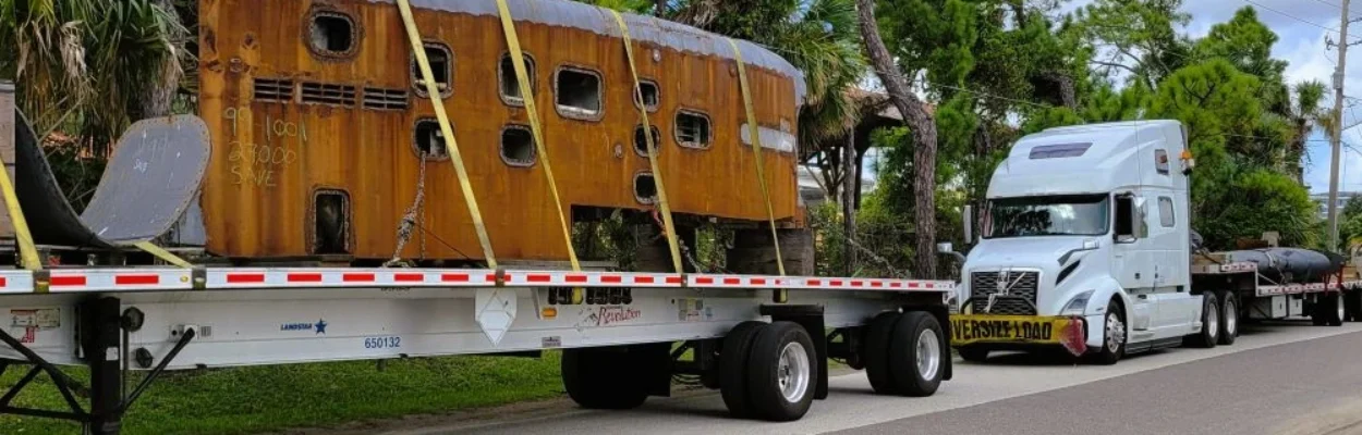 A section of the USS Jacksonville sits on a flatbed trailer with yellow straps holding it in place.