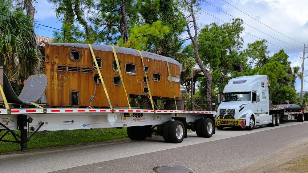 A section of the USS Jacksonville sits on a flatbed trailer with yellow straps holding it in place.