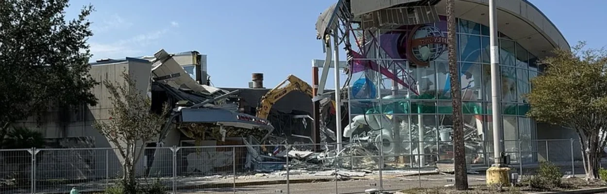 A crane is shown tearing down a mall building surrounded by a chain-link fence.