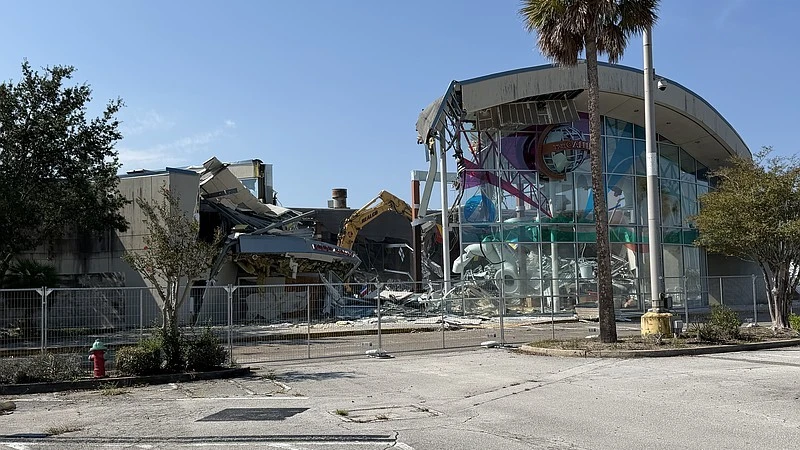 A crane is shown tearing down a mall building surrounded by a chain-link fence.