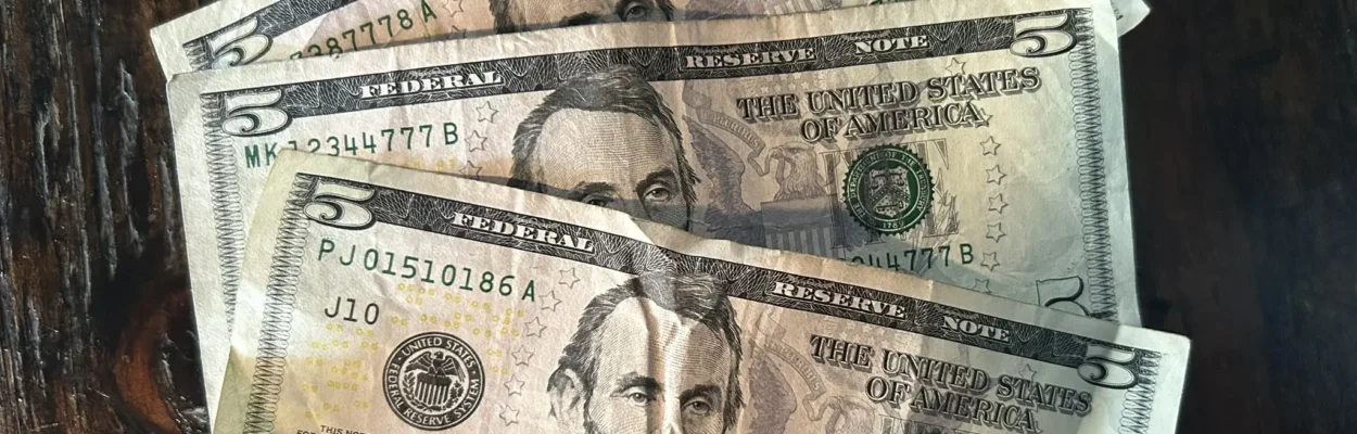 Three U.S. five-dollar bills are spread out on a dark wooden table.