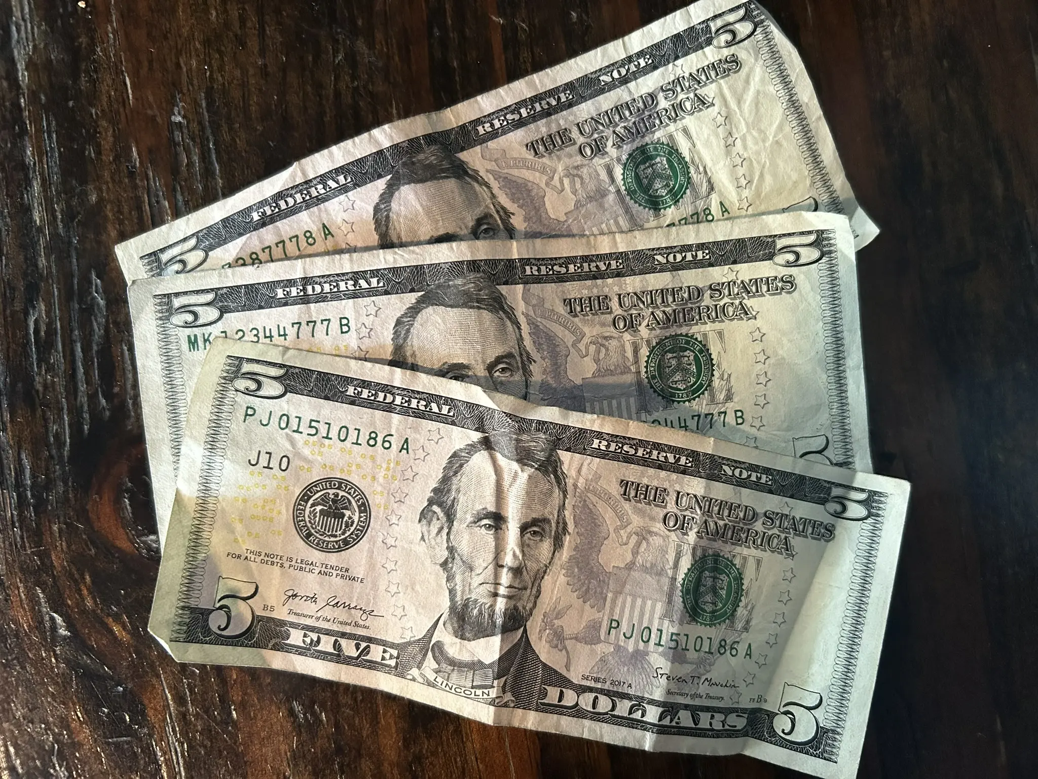 Three U.S. five-dollar bills are spread out on a dark wooden table.