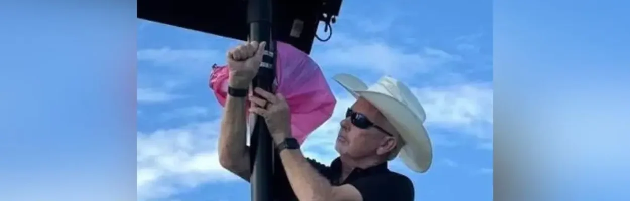 A man in a black shirt and white cowboy hat is taking a pink plastic bag and covering part of a Flock Camera.