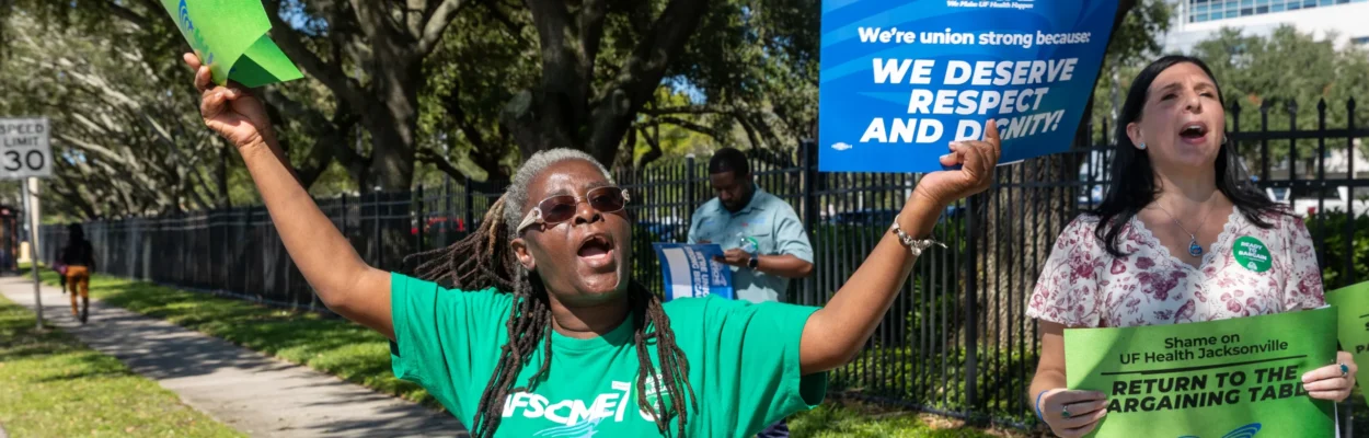 AFSCME Local 1328 President Mary Blocker-Williams wears a green T-shirt with a green sign in her right hand and blue sign in her left hand demanding UF Health Jacksonville return to the negotiating table.