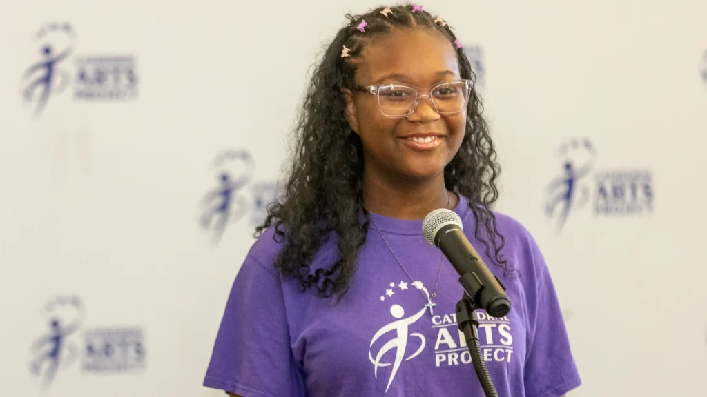 A’laiya Crew wears a purple Cathedral Arts Project T-shirt as she speaks behind a microphone
